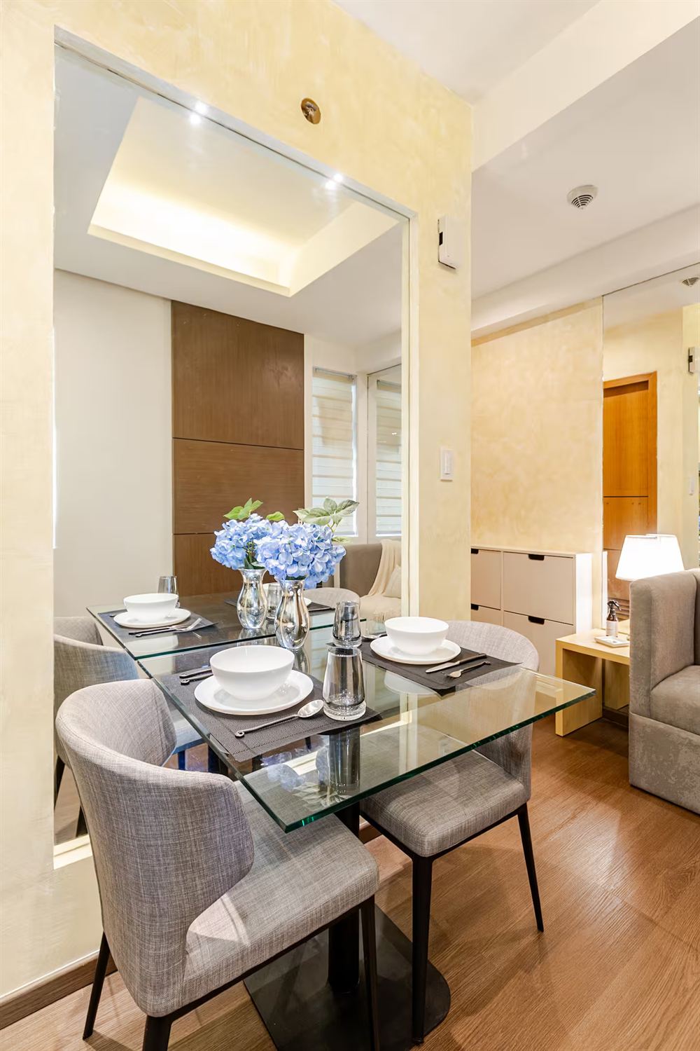 Dining area with a glass table and four grey linen chairs beside a full-height mirror